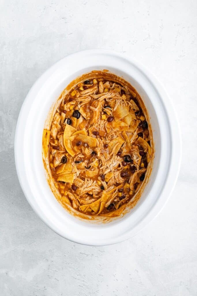 Overhead shot of shredded chicken, black beans, and tortilla chips in creamy sauce inside a white slow cooker.
