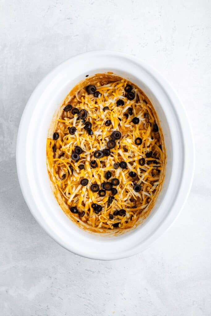 Baked casserole in an oval white dish, topped with shredded cheese and black olives on a light surface.