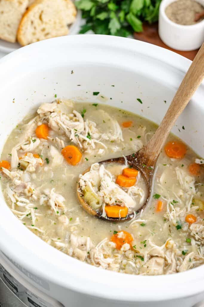 Slow cooker with chicken, barley, and vegetable soup, a wooden ladle, and sliced bread in the background.