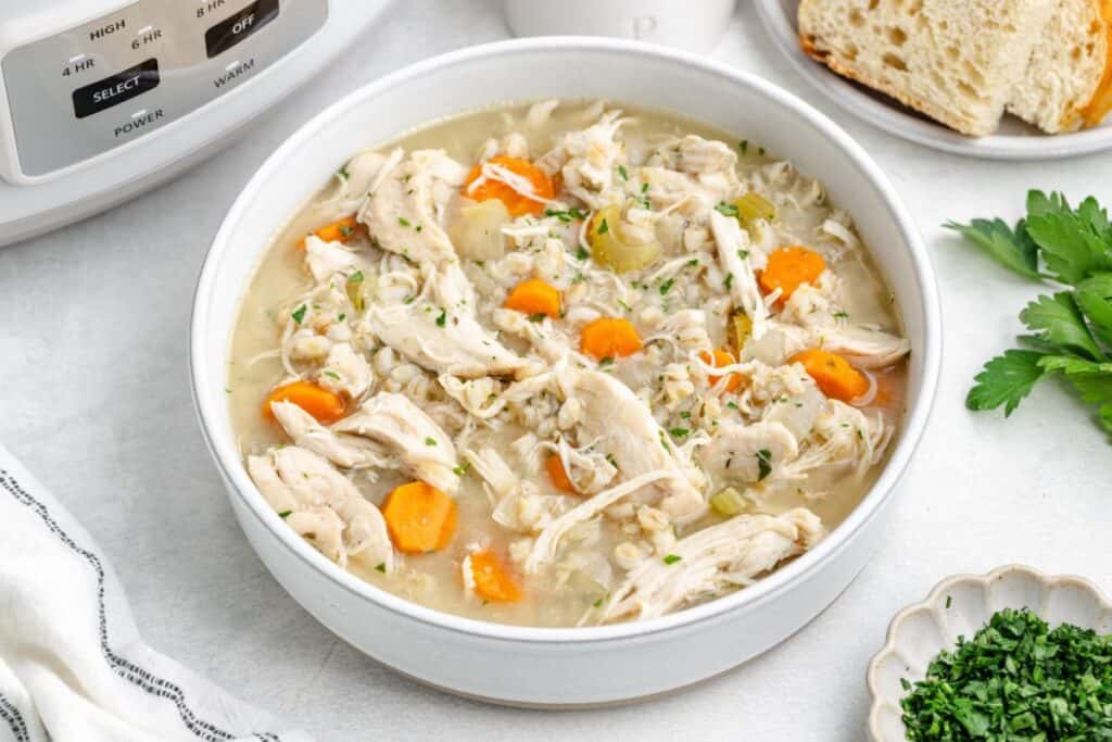 Chicken and barley soup with carrots and herbs in a bowl, next to sliced bread and fresh parsley.