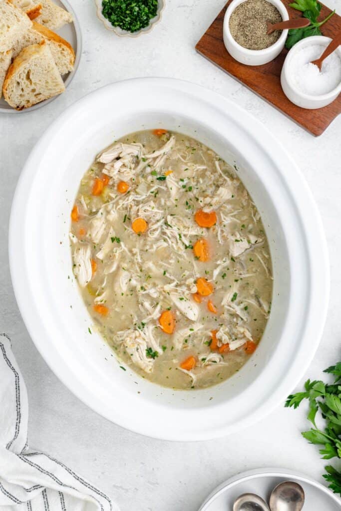 Oval slow cooker with shredded chicken, carrots, and broth, garnished with herbs. Bread and seasonings nearby.