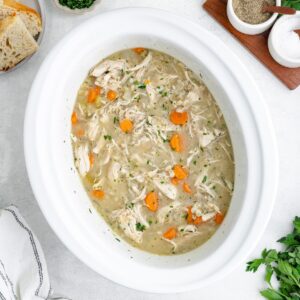 Crockpot chicken barley soup in a white slow cooker, with bread and herbs nearby.