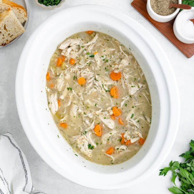 Crockpot chicken barley soup in a white slow cooker, with bread and herbs nearby.