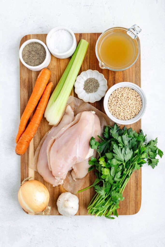 Ingredients on a wooden board: raw chicken breasts, vegetables, seasonings, pearl barley, and chicken broth.