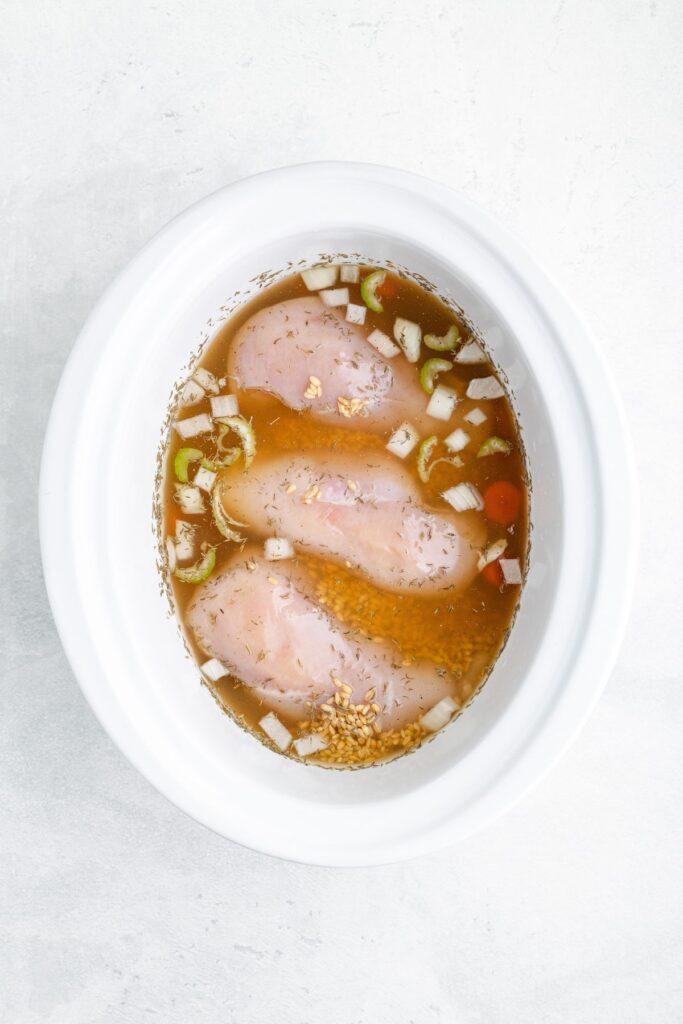 Raw chicken breasts, chopped onions, and vegetables in broth inside a white slow cooker on a light surface.