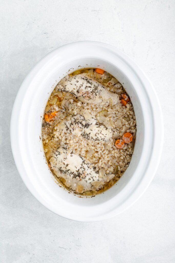 Chicken, rice, carrots, and herbs in a white slow cooker, viewed from above on a light surface.