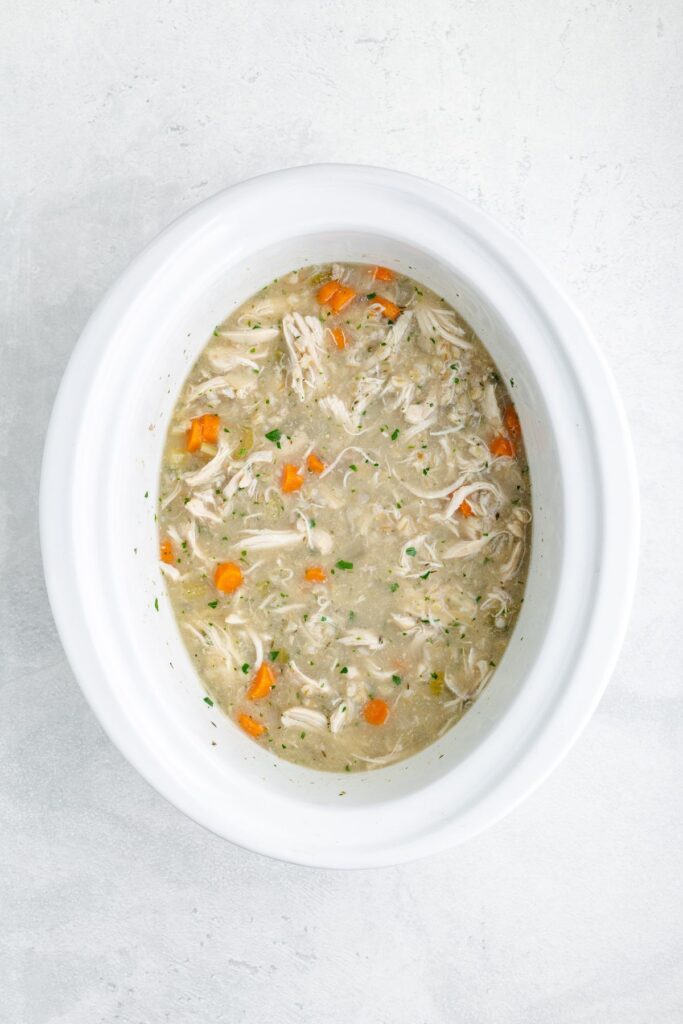 Oval slow cooker with shredded chicken soup, carrots, herbs, and broth on a light surface.