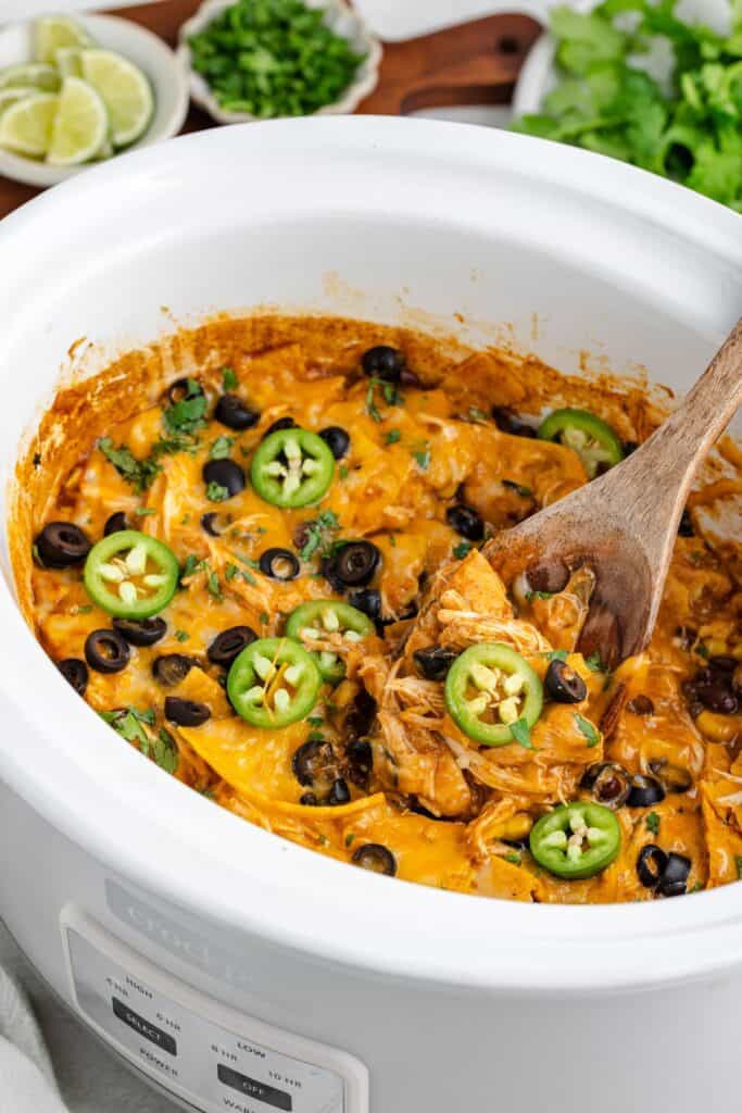 Slow Cooker Chicken Enchilada Casserole in a white slow cooker with a wooden spoon.