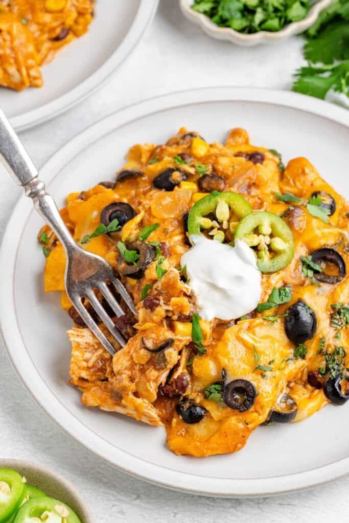 Cheesy enchilada casserole topped with beans, olives, corn, and sour cream; fork cutting into it.