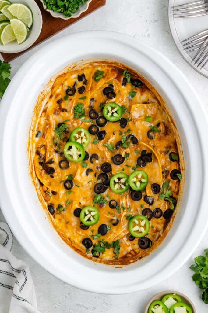 Crockpot chicken enchilada casserole with cheese, black olives, jalapeƱos, and cilantro in an oval dish; plates and limes nearby.
