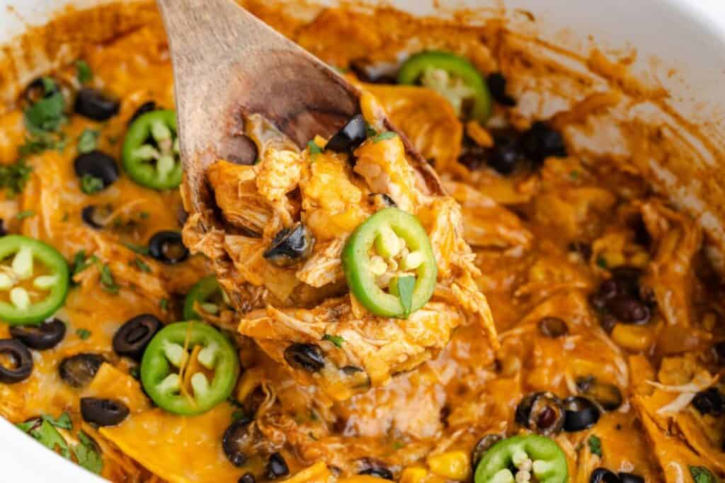 Wooden spoon scooping cheesy chicken enchilada casserole with black olives, corn, and jalapeƱos from a crock pot.