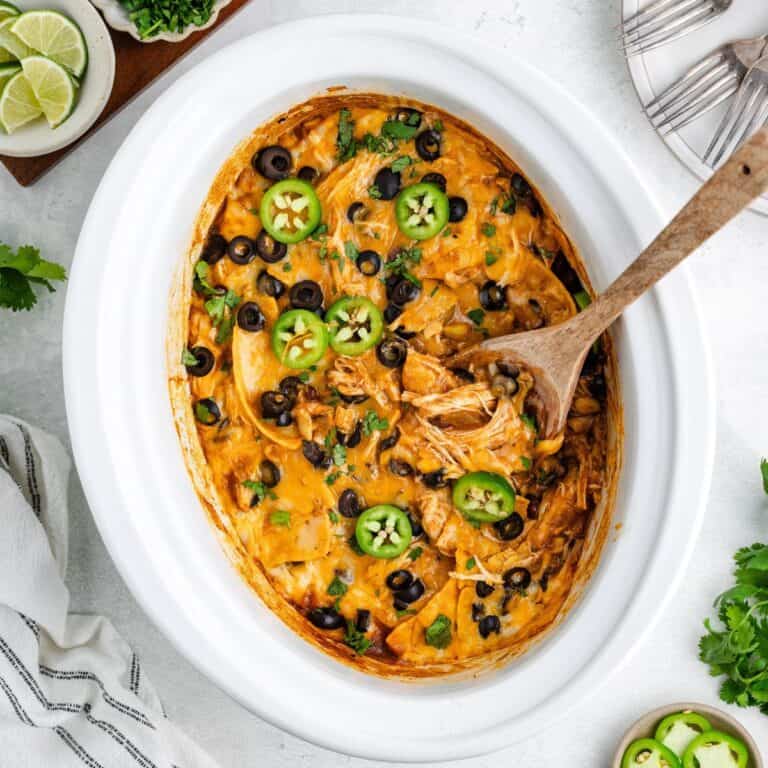 Slow Cooker Chicken Enchilada Casserole topped with olives, jalapeños, cilantro; wooden spoon inside.