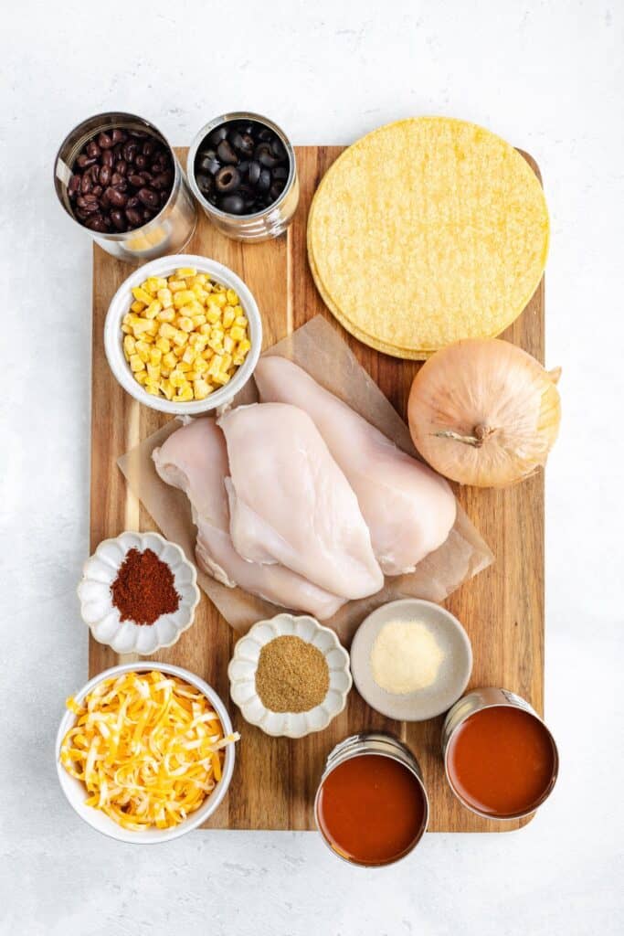 Wooden board with raw chicken, tortillas, onion, cheese, spices, corn, beans, olives, and enchilada sauce.