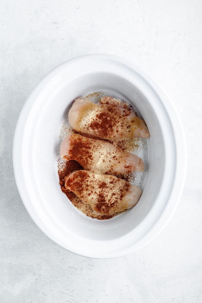 Three spiced raw chicken breasts sit in a white slow cooker on a light-colored surface.
