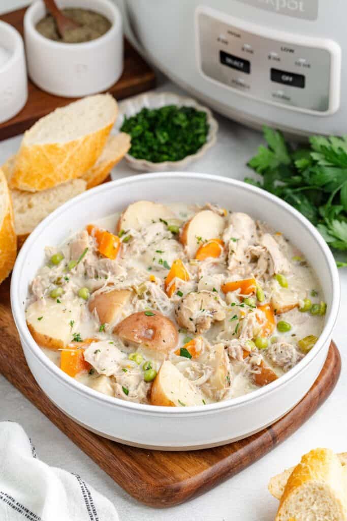 Creamy crockpot chicken stew with potatoes and carrots in a bowl, garnished with herbs, beside sliced bread and parsley.
