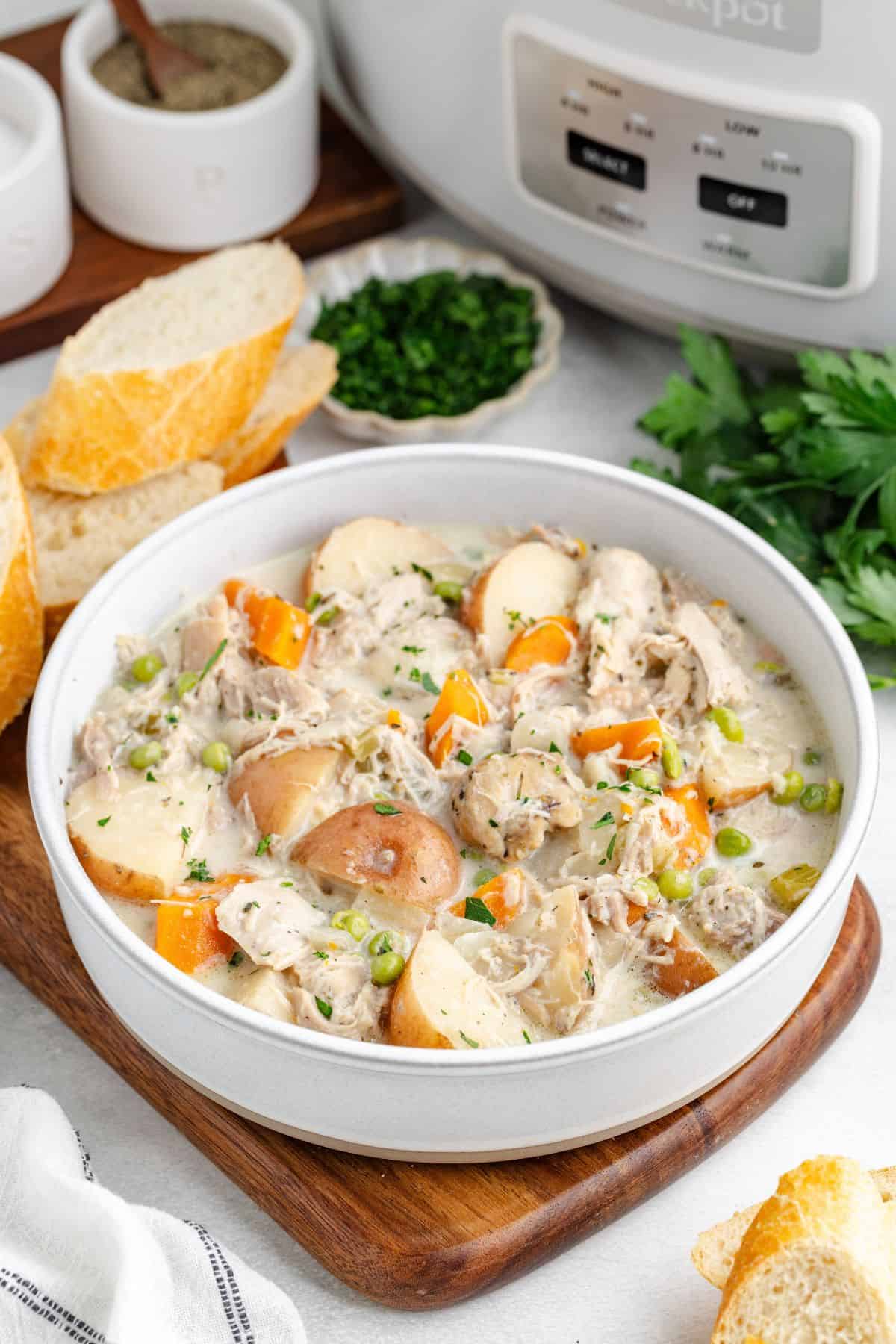 Slow Cooker Chicken Stew Chicken Stew in a white bowl on a cutting board with sliced bread. Behind the bowl is a white slow cooker and fresh herbs.