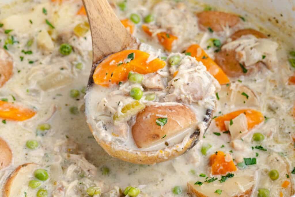 Wooden ladle holding creamy chicken stew with vegetables above a slow cooker of the same dish.
