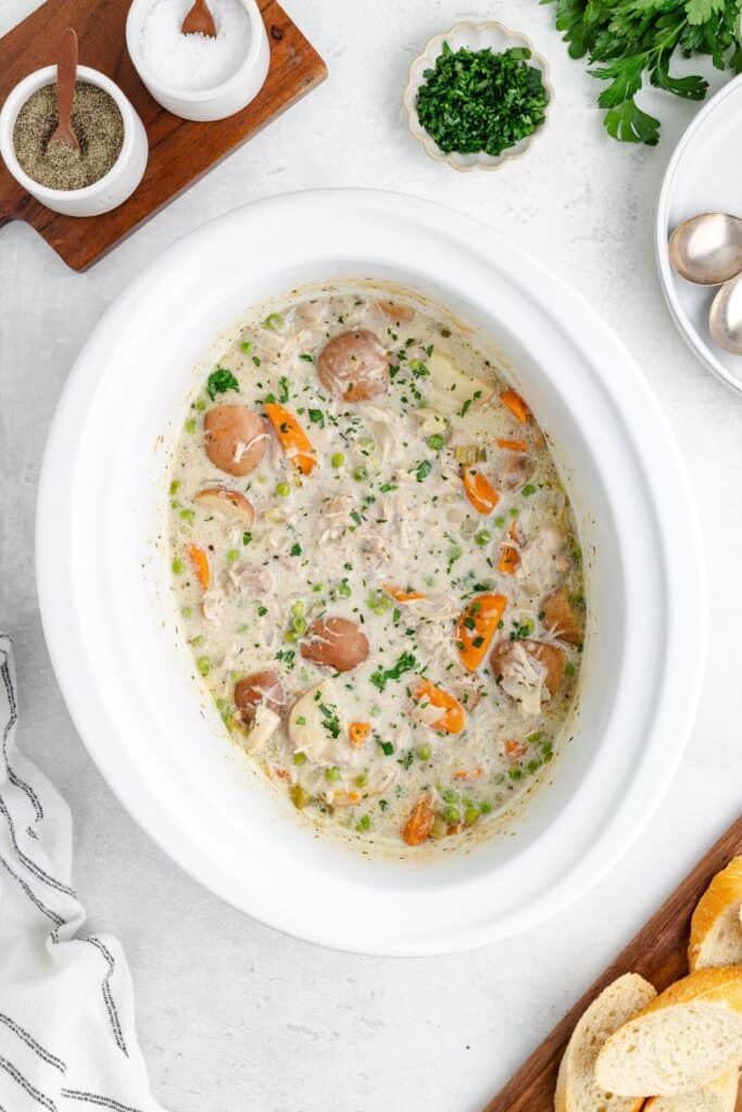 Slow cooker chicken stew, carrots, peas, potatoes, and herbs on a white surface.