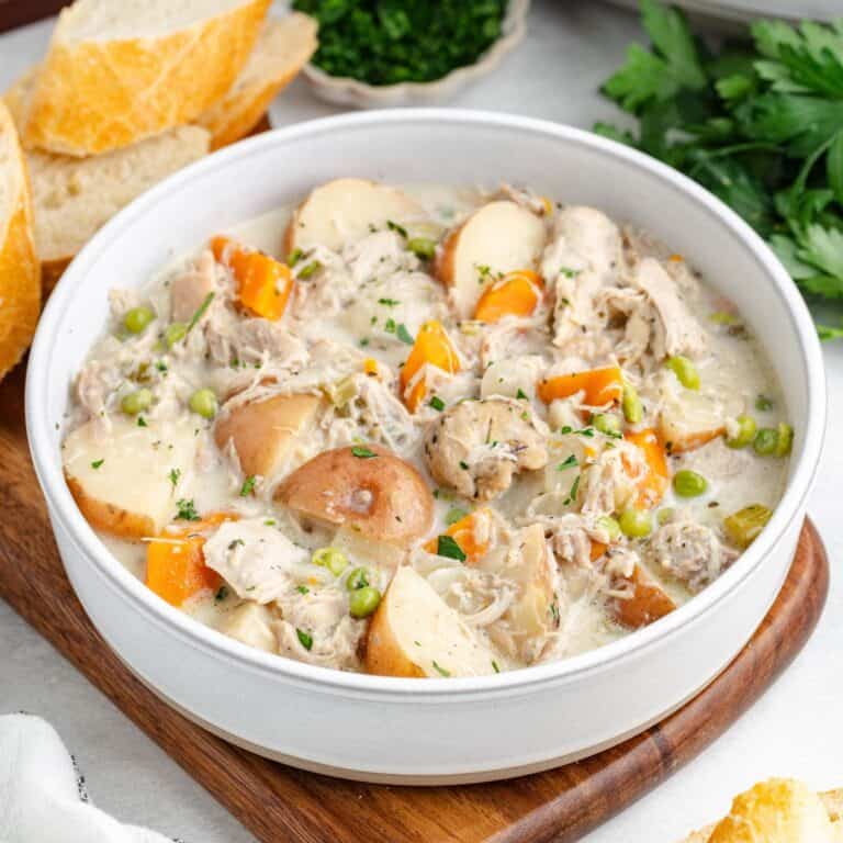 Creamy slow cooker chicken stew with vegetables in a bowl on a wooden board, bread slices beside it.