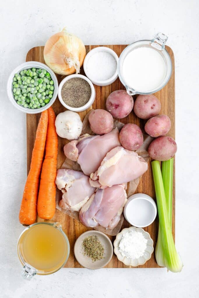 Raw chicken thighs, vegetables, seasonings, milk, broth, and flour displayed on a wooden board.