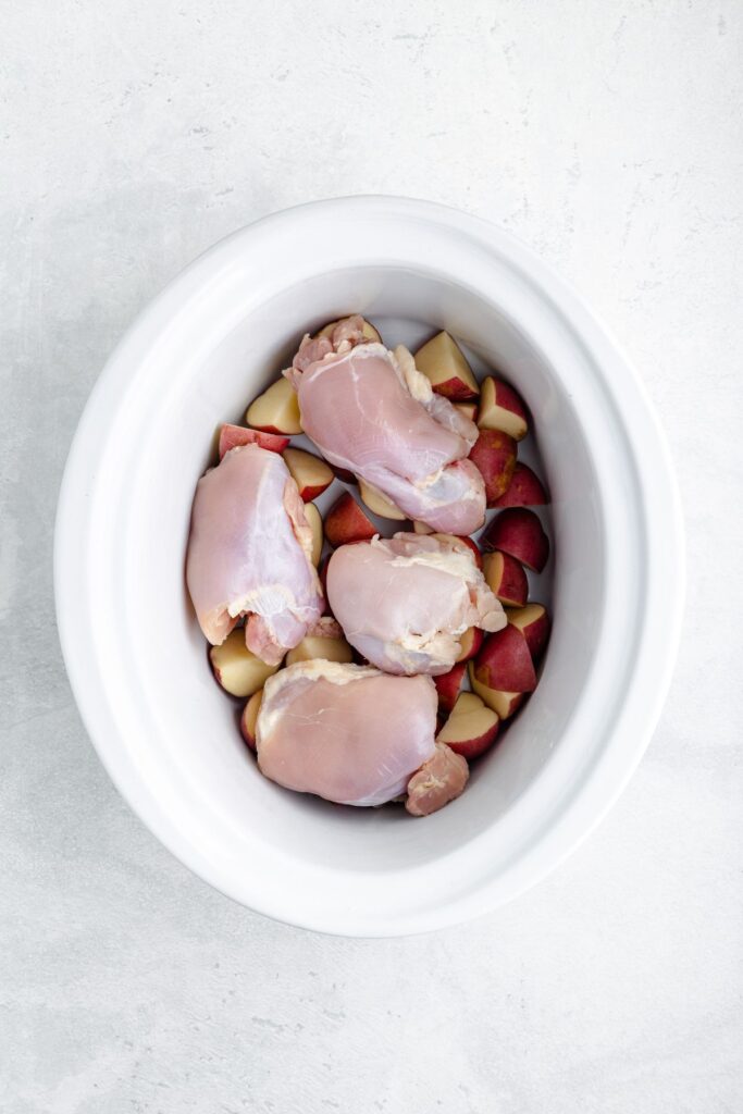 Raw chicken thighs atop chopped red potatoes in a white slow cooker on a light background.