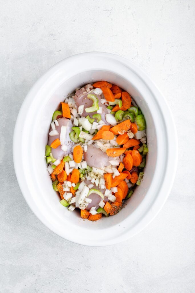 Raw chicken, carrots, celery, onions, and seasonings in a white slow cooker on a light surface.