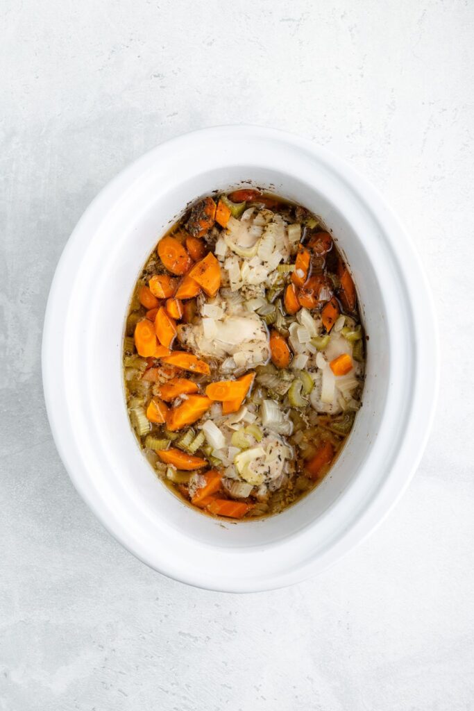 White slow cooker containing chopped carrots, onions, celery, and cooked chicken pieces in broth.