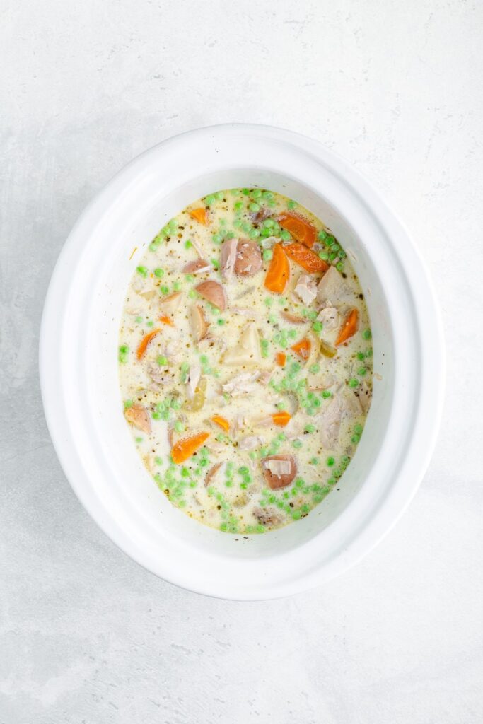 Creamy stew with peas, carrots, potatoes, and meat inside a white slow cooker, top-down view.