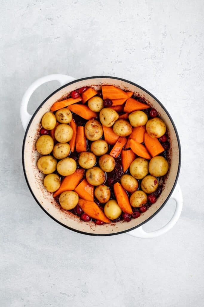 Baby potatoes, carrot wedges, and cranberries in a white enameled cast iron pot.