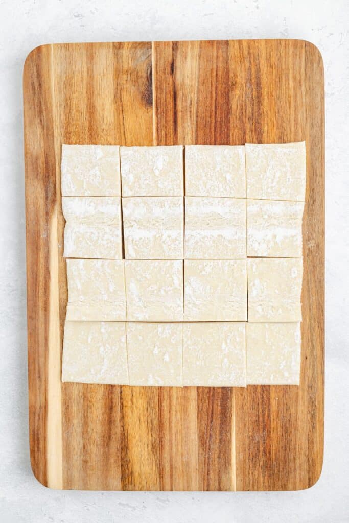 Sixteen flour-dusted raw dough squares arranged in a grid on a wooden board.