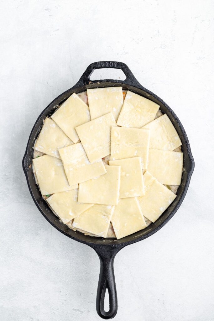 A cast iron skillet filled with savory filling topped with overlapping squares of raw pastry dough.