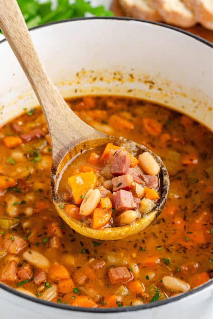 A wooden spoon lifts ham and bean soup with carrots from a large pot.