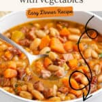 A spoon lifting ham and bean soup with vegetables from a white bowl.