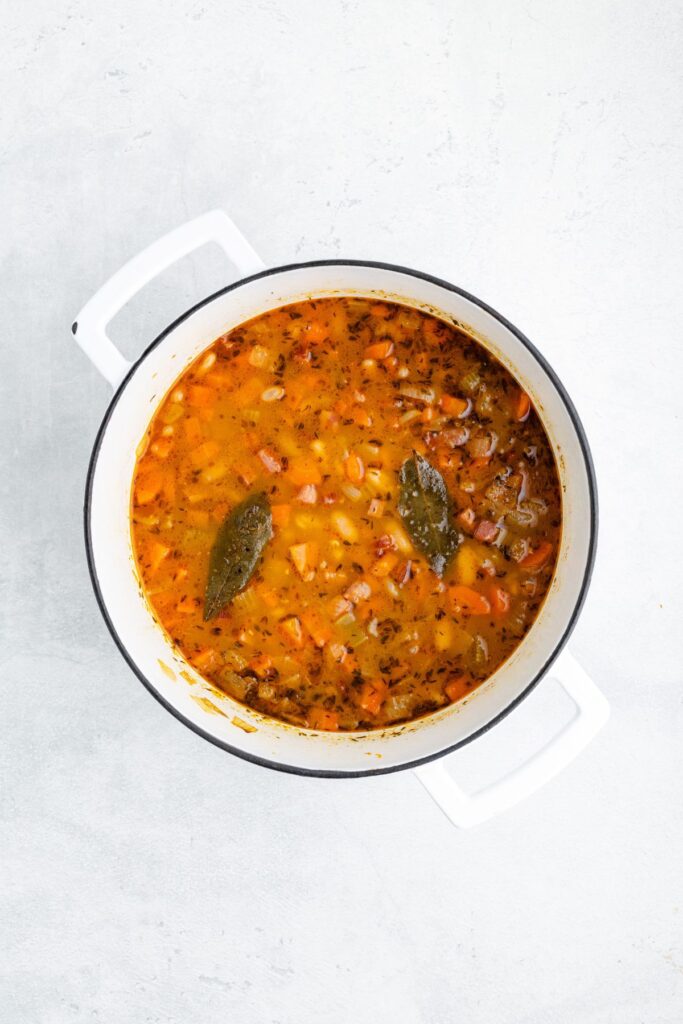 Ham, vegetable and bean soup with two bay leaves in a white pot.