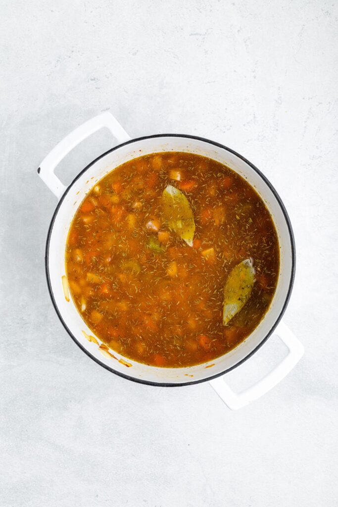 Top-down view of ham and vegetable soup with bay leaves in a white pot.