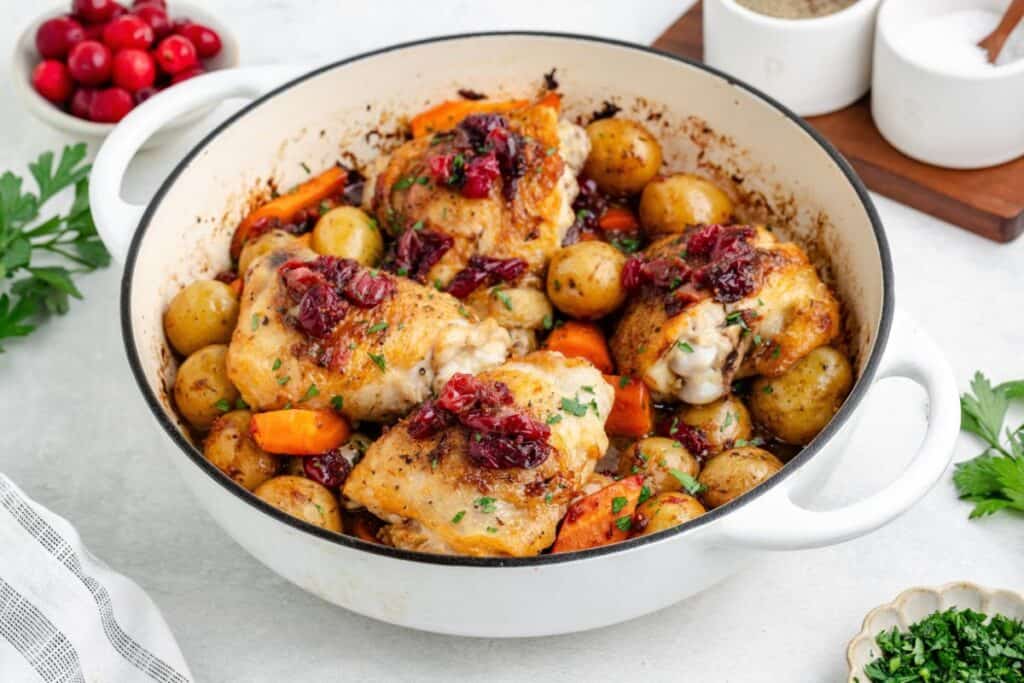 Roasted cranberry chicken thighs,  carrots, and potatoes in a white enameled cast-iron pan.