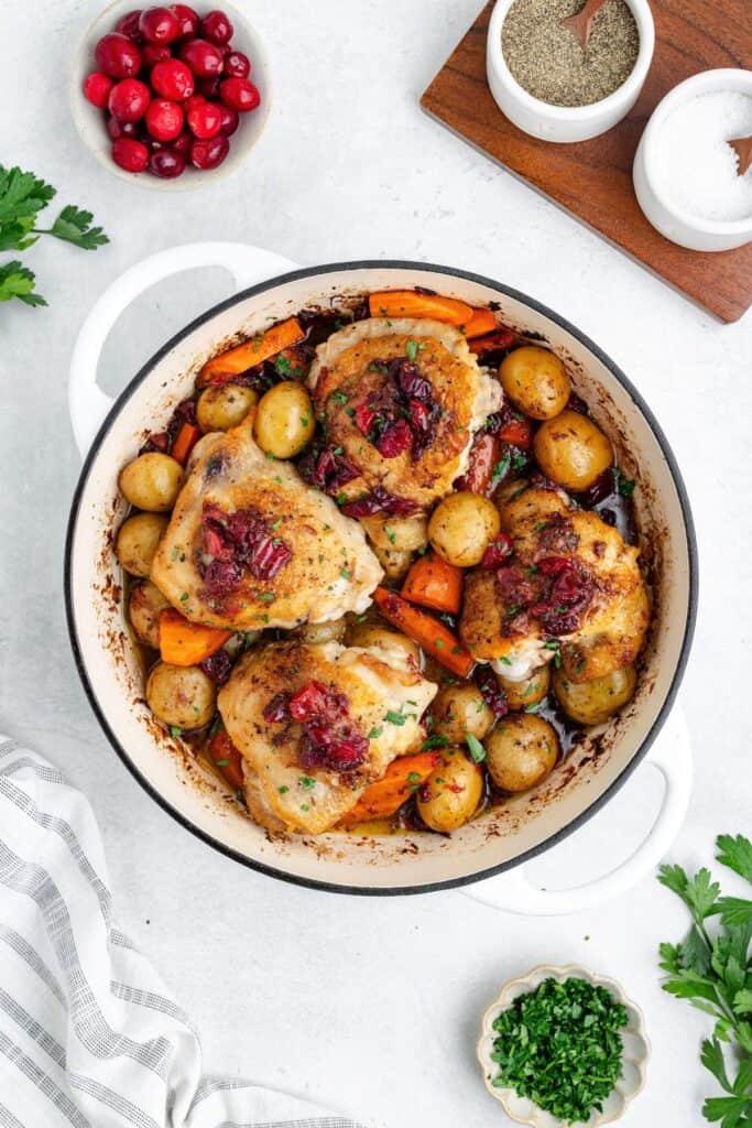 Roasted chicken thighs with potatoes, carrots, and cranberries in a white enameled cast iron pot.