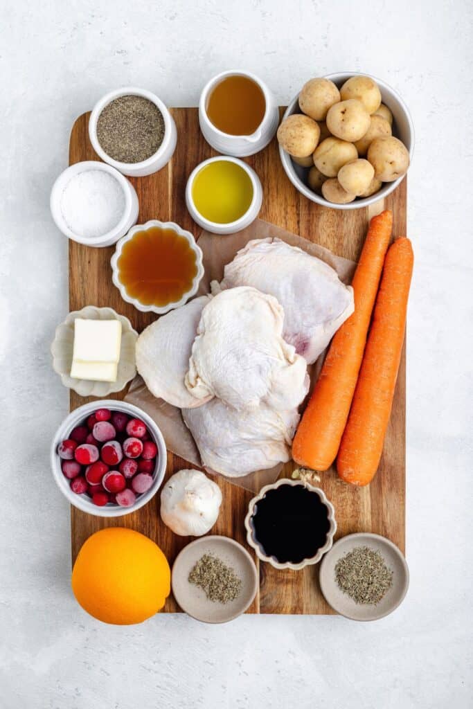 Raw chicken thighs with carrots, potatoes, cranberries, orange, garlic, butter, oils, and seasonings on a board.